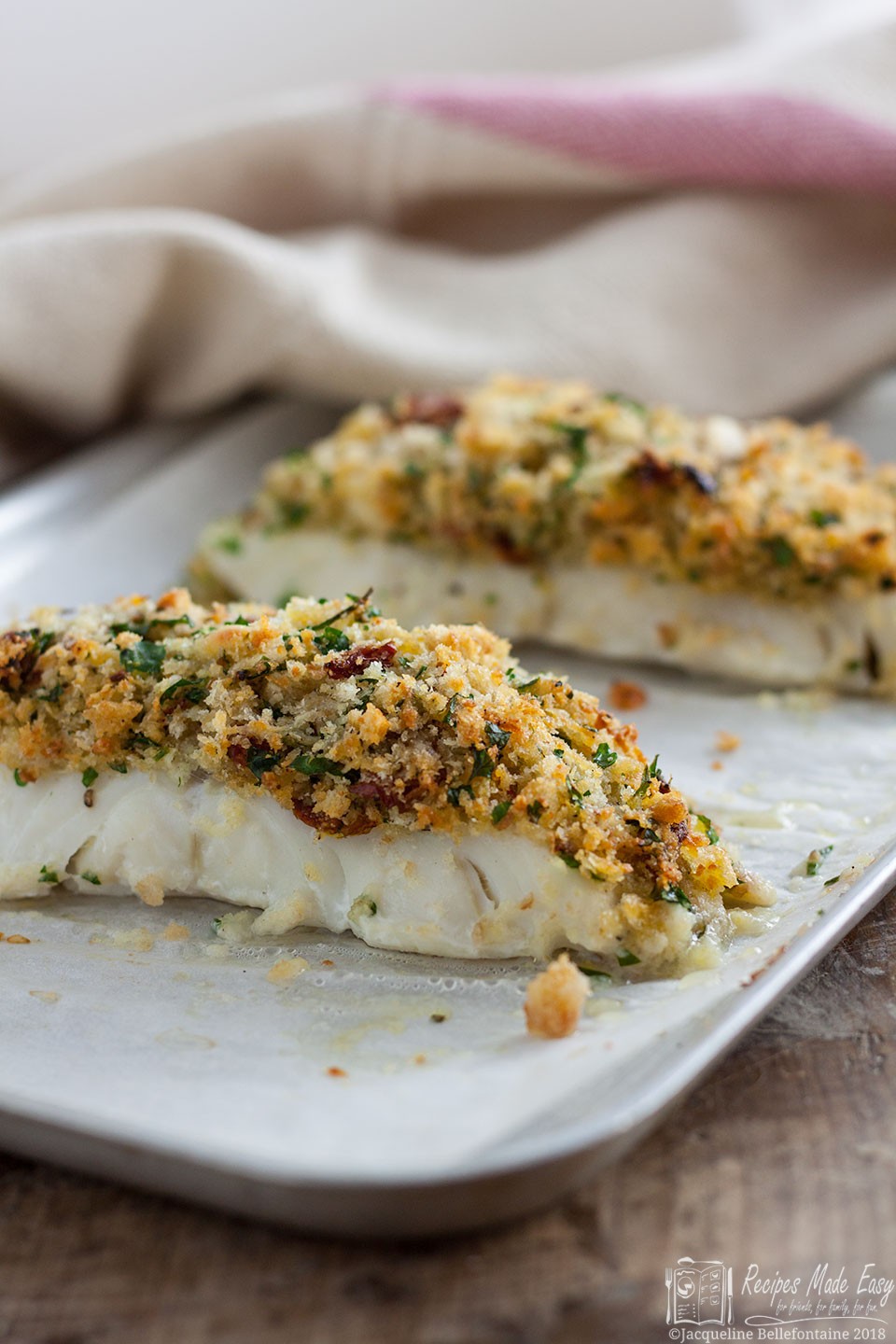 Two Herb crusted roast cod pieces on a baking tray with oven cloth in background