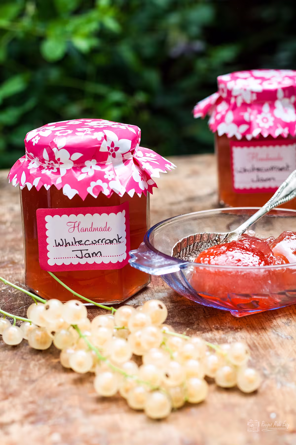 2 jars of whitecurrant jam with some jam in a glass bowl and fresh whitecurrants in front.