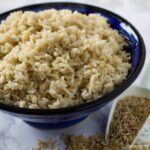 Bowl of cooked brown rice with scoop of uncooked brown rice grains spilling out beside the bowl.