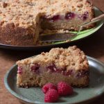 slice of raspberry crumble cake on plate with fresh raspberries. whole cake behind.