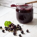 open jar of blackcurrant jam with knife resting on top. Fresh blackcurrants on worksurface in front.