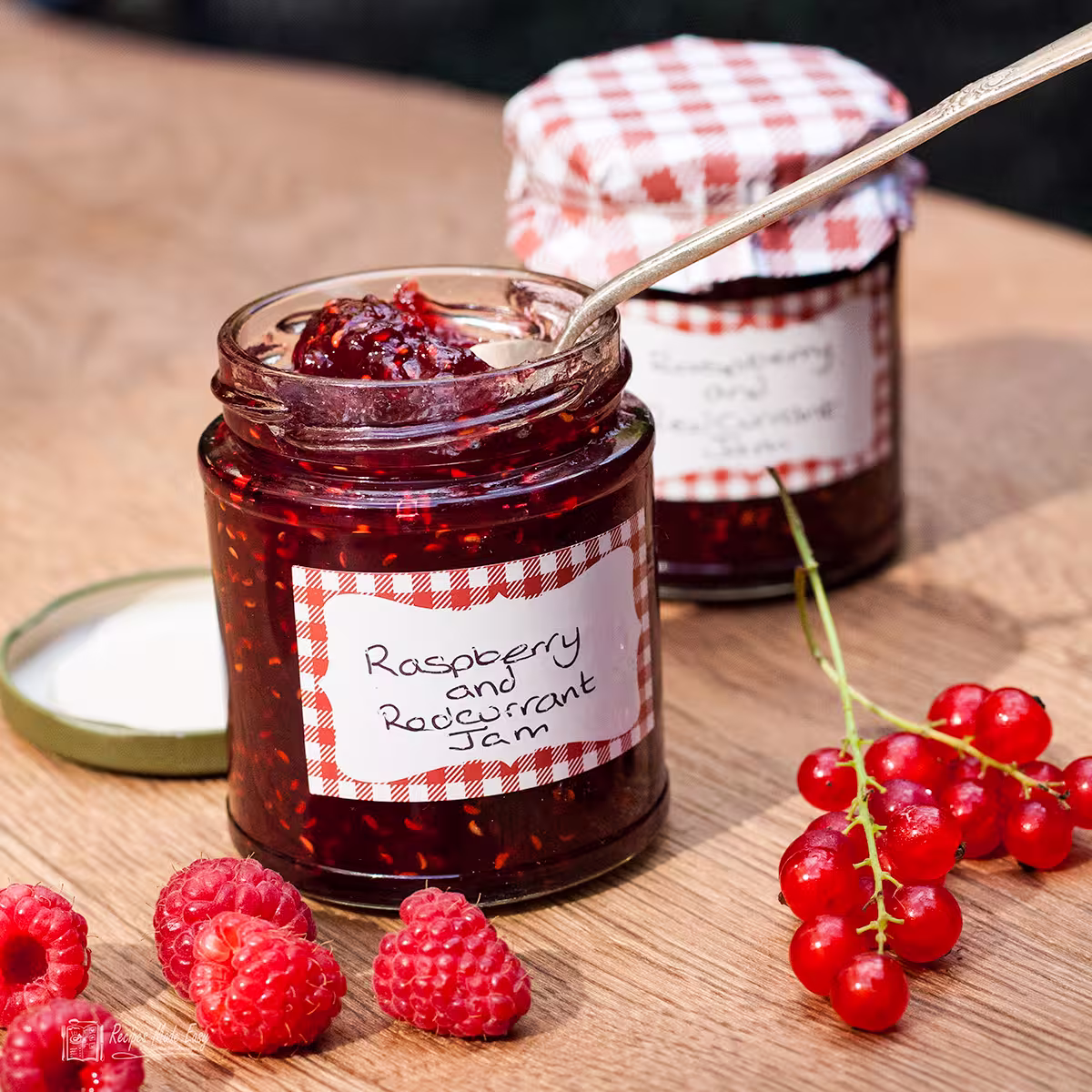 two jars of raspberry and redcurrant jam. one open with spoon. fresh raspberries and redcurrants in front of jars.