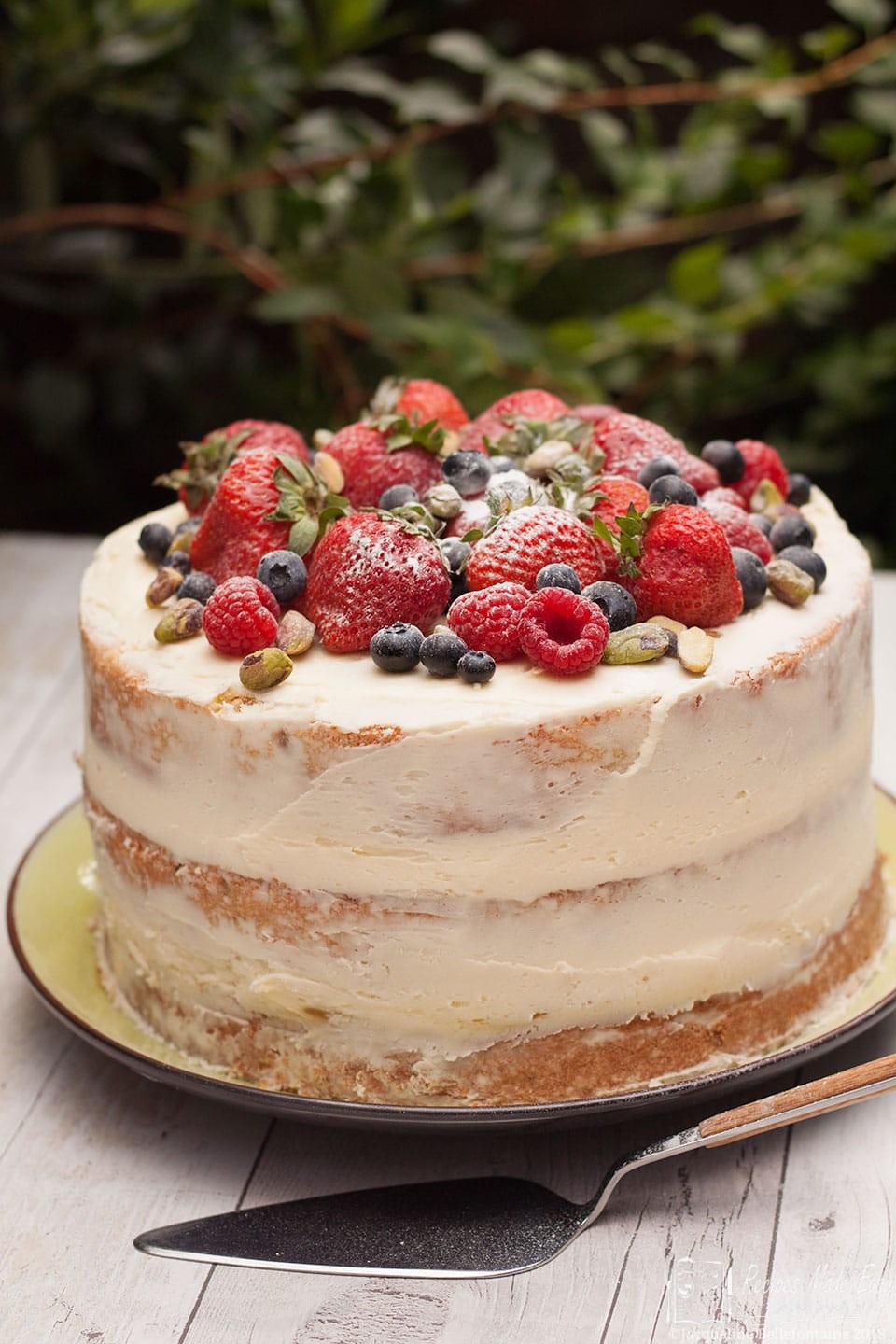 whole pistachio and lime cake decorated with summer berries with a cake slice in front.