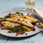 plate of sea bass with peas, bacon and potato with fish knife and fork on side. wine glass behind